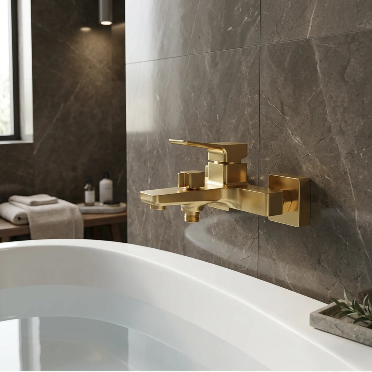 A modern gold wall-mounted bathtub faucet installed on a dark gray marble-tiled wall above a white bathtub.