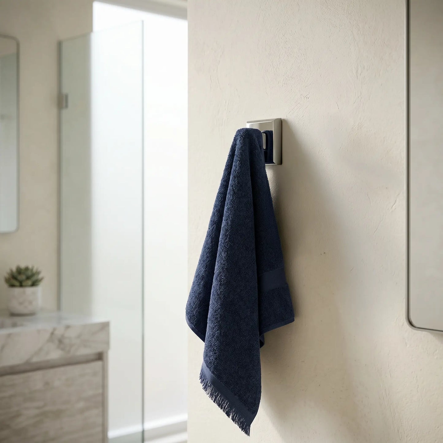 A minimalist bathroom setting showing the Kludi Rak chrome robe hook mounted on a neutral wall with a navy blue towel hanging from it, featuring a marble vanity and glass shower in the background.
