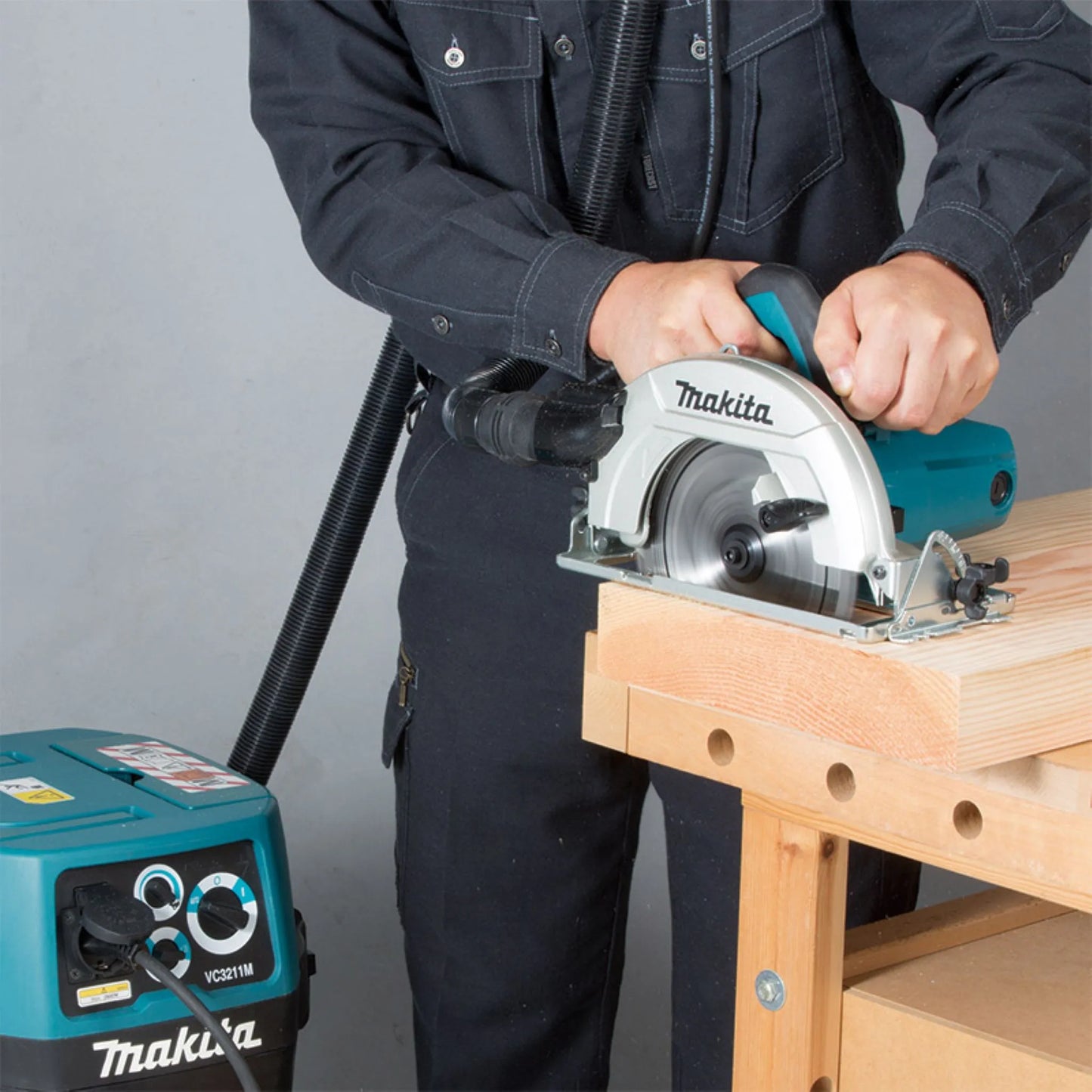 Professional carpenter using a Makita corded circular saw with a dust extraction vacuum attachment to cut a thick timber plank on a workbench.