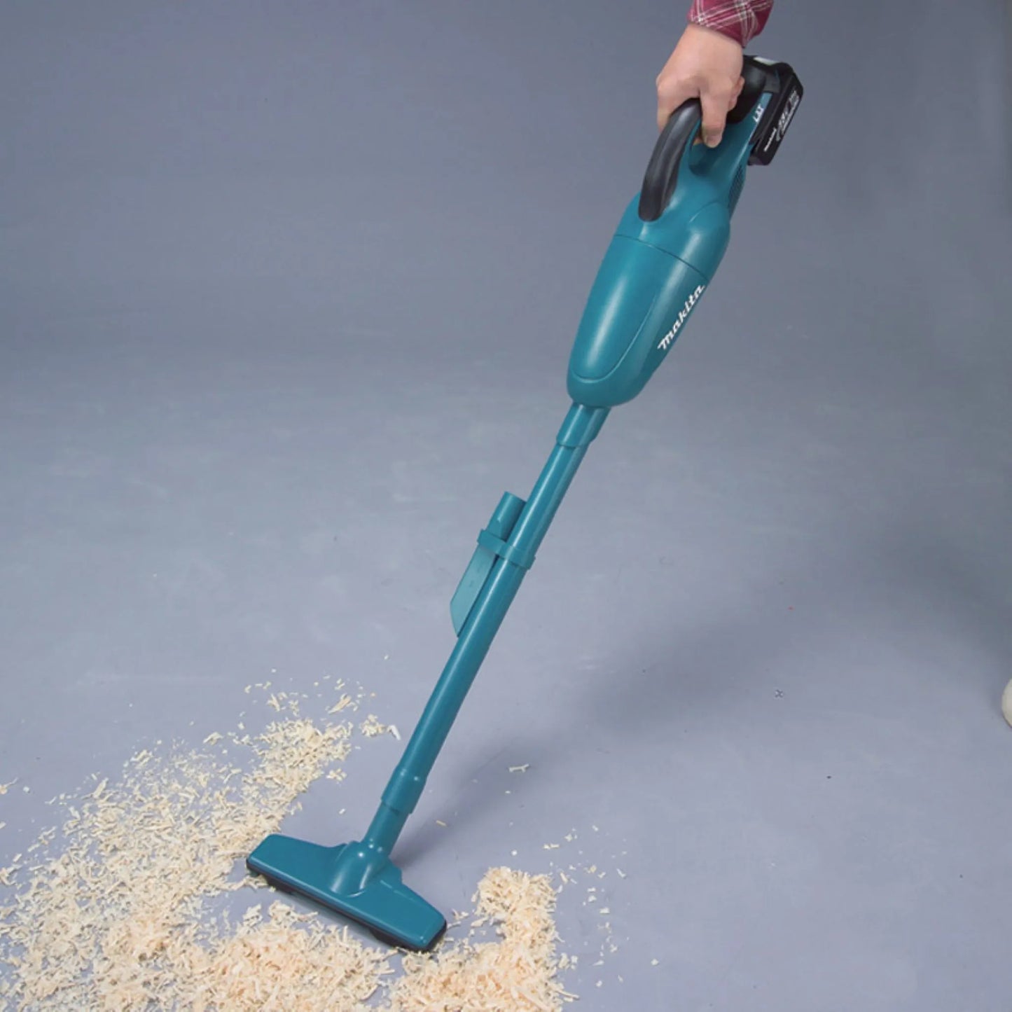 Person using a Makita 18V LXT cordless stick vacuum with a floor nozzle attachment to clean wood shavings off a floor.